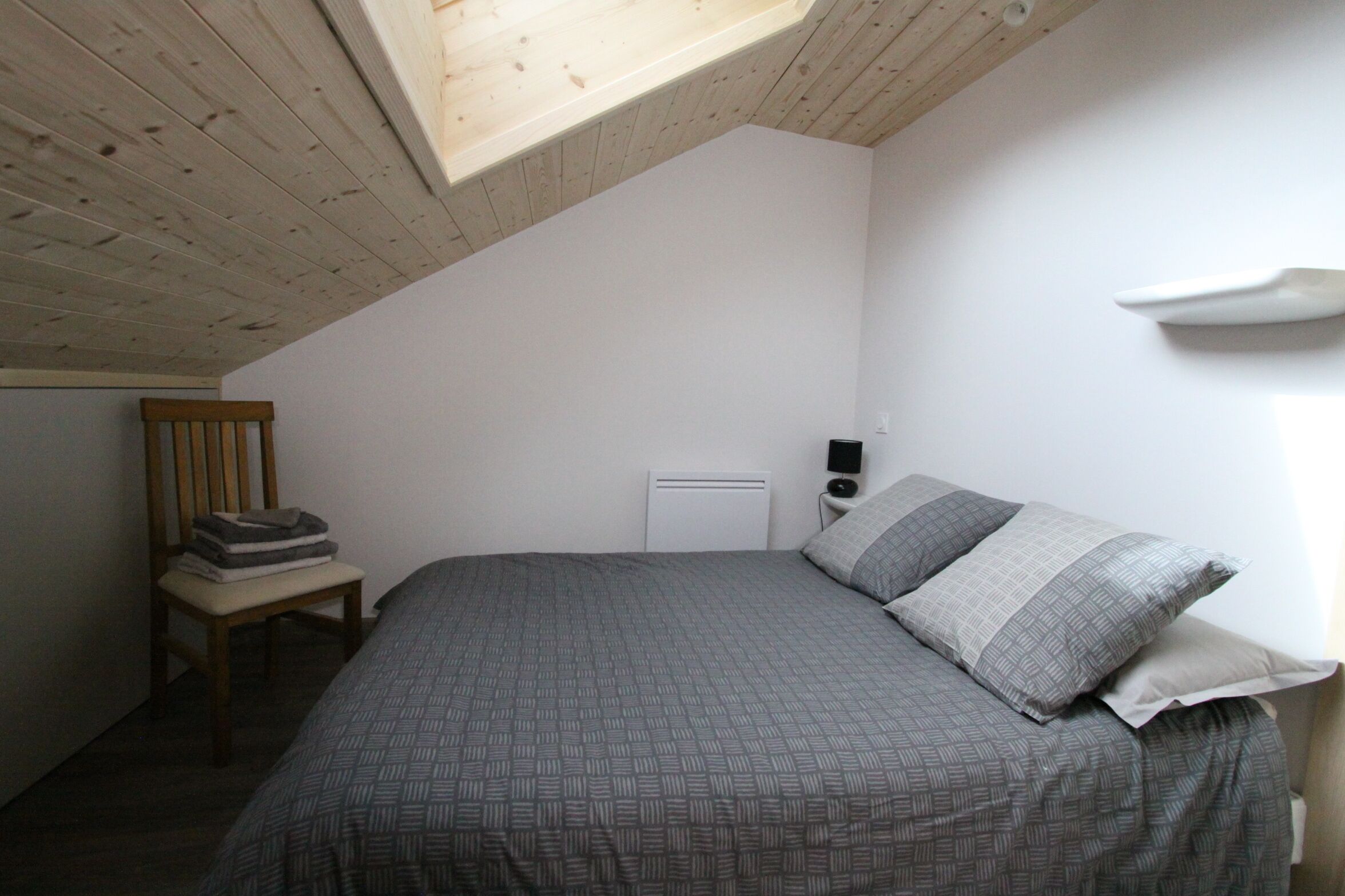 Een slaapkamer met een schuine kap, een bed met grijze beddengoed, een stoel met opgevouwen dekens en een witte radiator.