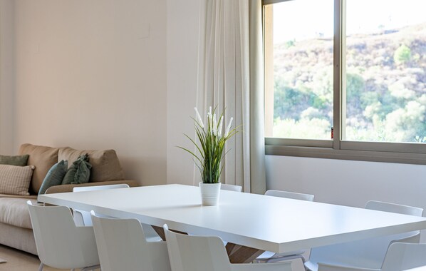 Ein modernes Wohnzimmer mit Esstisch in Weiß, weißen Stühlen, einem beige farbenen Sofa mit Kissen und einer Topfpflanze.