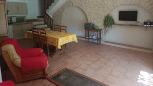 A dining area with a table set for four, a wooden chair with a red cushion, a small table with a basket and books, and a stone wall with a mounted TV.