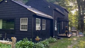 A black wooden house with a porch, surrounded by lush greenery and a stone pathway.
