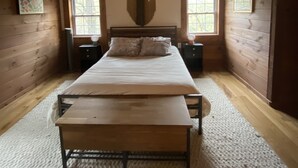 A bedroom with a wooden ceiling, a bed with pillows, a bench, two nightstands, and a wall-mounted light fixture.