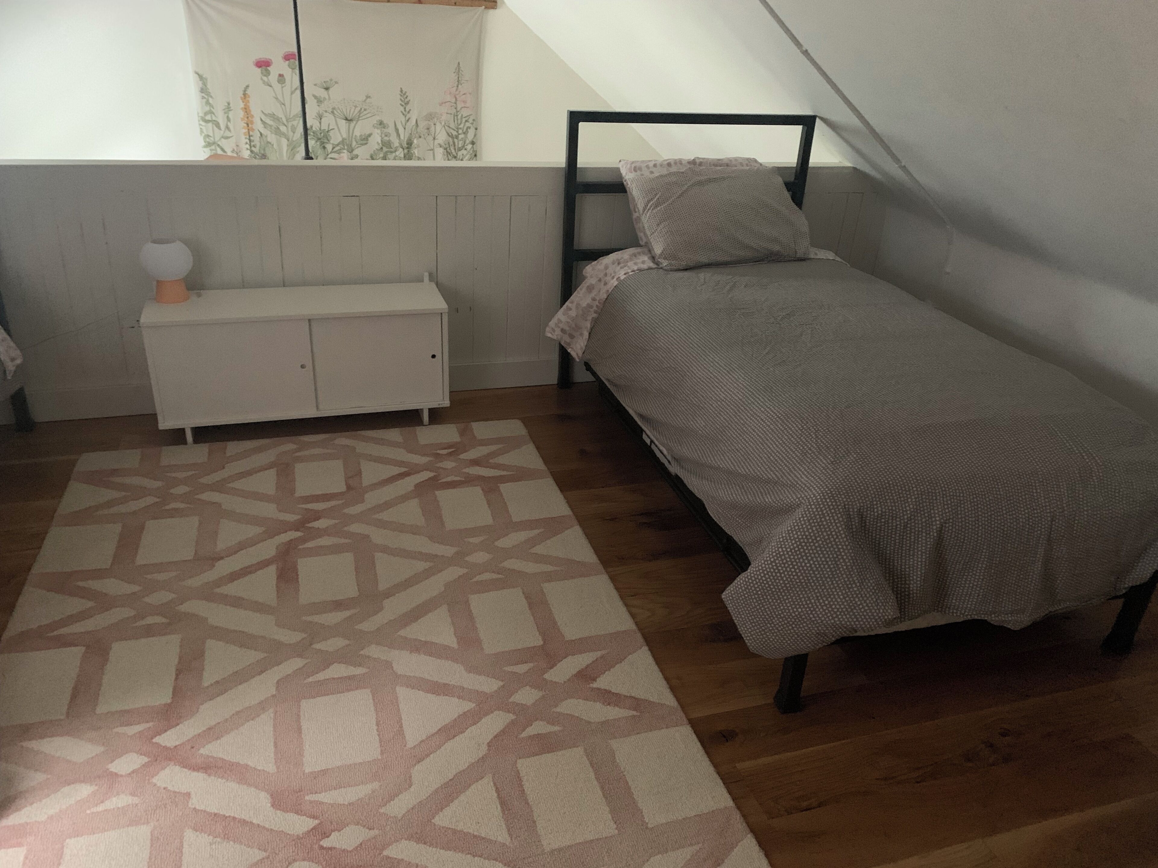 A small, single-bed room with a sloped ceiling, a patterned rug, a white dresser, and a window with a floral curtain.