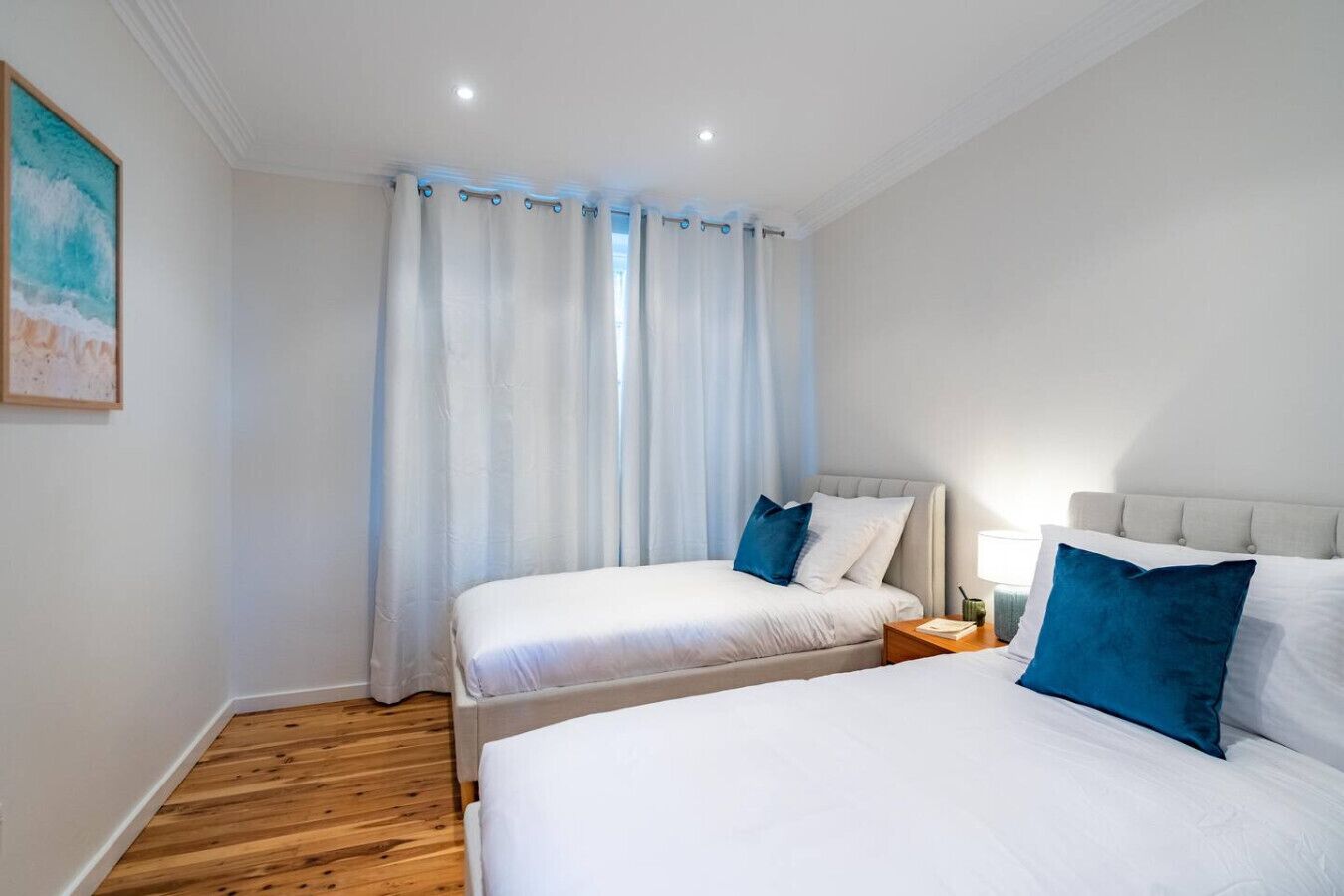 A bedroom with two beds, a wooden floor, a painting on the wall, and a window with curtains.