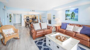 Amplia sala de estar con un sofá de cuero marrón, un sillón de mimbre y una mesa de centro con objetos decorativos. Se ve una cocina al fondo, y un cuadro con temática de playa adorna la pared.