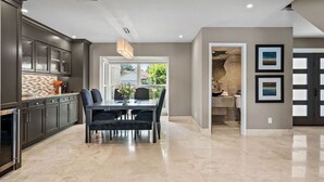 A modern kitchen with dark cabinets, a dining area with a table and chairs, and a view of the outdoors through a large window.
