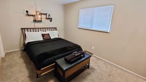A bedroom with a bed, a bedside table, a lamp, and a window with blinds. - Stylish Steampunk Home for Extended Stays (Denver)
