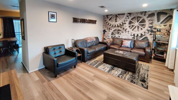 A modern living room with a leather sofa, a blue armchair, and a coffee table. There is a wall with a large gear decoration and a framed picture. - Stylish Steampunk Home for Extended Stays (Denver)