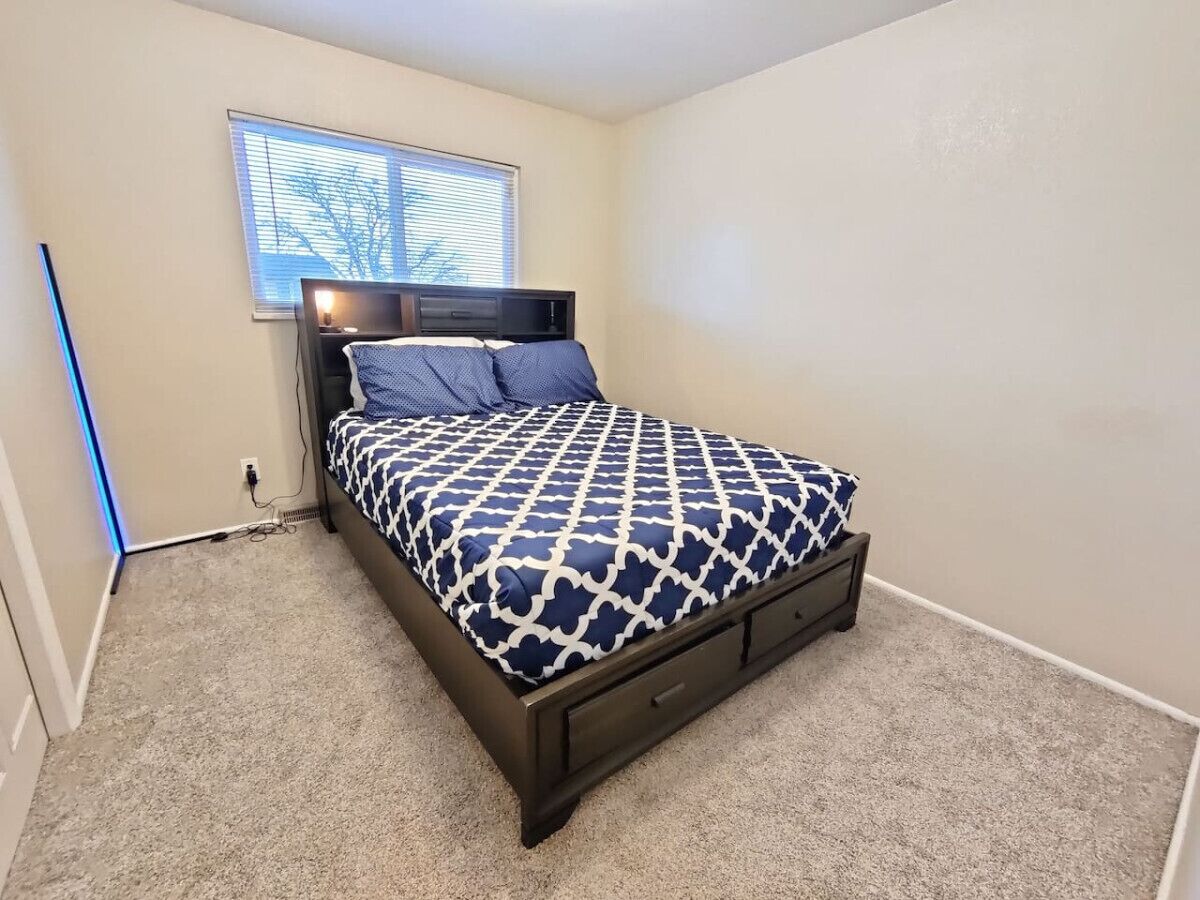 A bedroom with a bed, a nightstand, a window, and a wall socket.