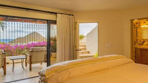 A bedroom with a large bed, a balcony with wicker furniture, and a view of the sea and mountains.