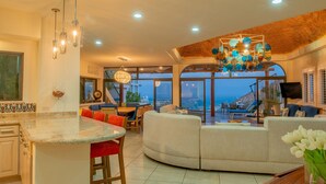 A modern kitchen with a central island, a dining area with a view, and a living room with a large sofa and a decorative ceiling light.