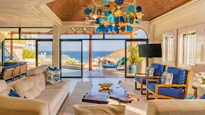 A spacious living area with a large window offering a view of the sea, a unique ceiling chandelier, and comfortable seating.