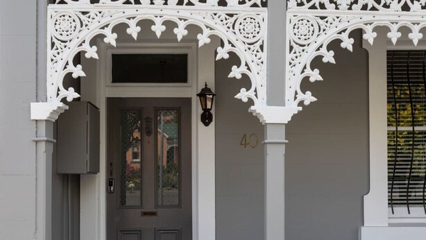 A grey building with a white ornate entrance, a black door, and a window with blinds.