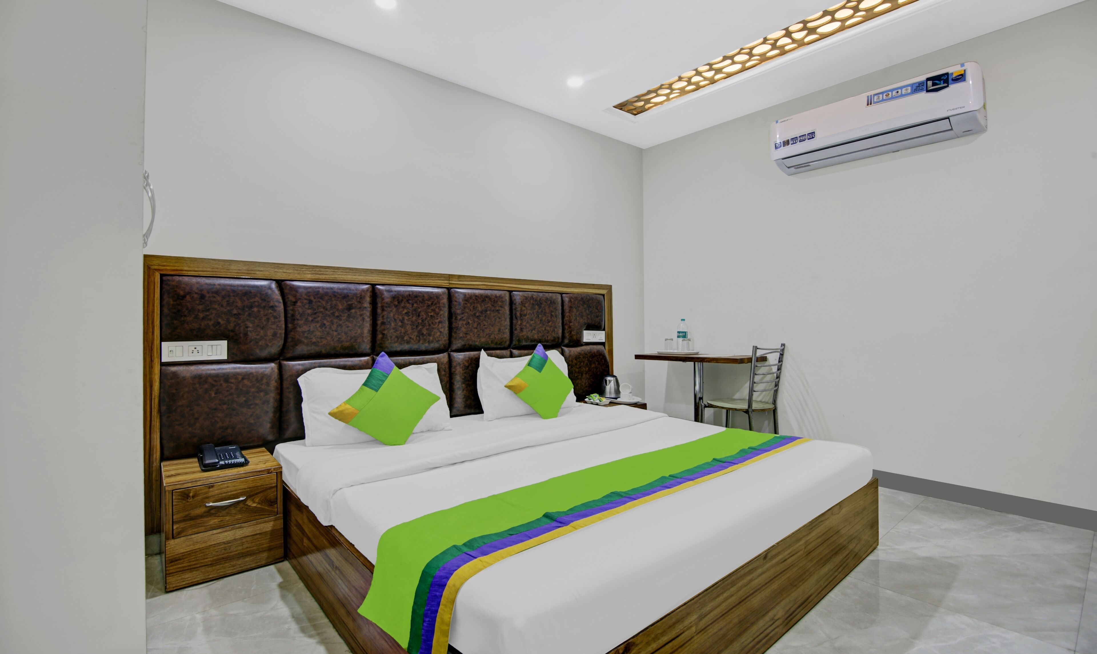 Habitación de hotel con cabecera de madera, cama con ropa blanca y almohadas de colores, mesita de noche con teléfono y botella de agua, y unidad de aire acondicionado en la pared.