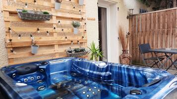Een hottub met een blauw marmerdesign, een houten muur met hangende lampen en potplanten, een houten omheining en een tafel met stoelen.