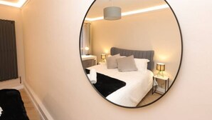 Une chambre à coucher avec un grand miroir, un lit recouvert de draps blancs, deux lampes de chevet et une tête de lit grise.