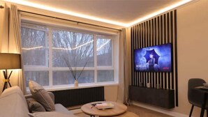 Un salon moderne avec un canapé, une table basse, un téléviseur fixé au mur et une fenêtre avec des rideaux.