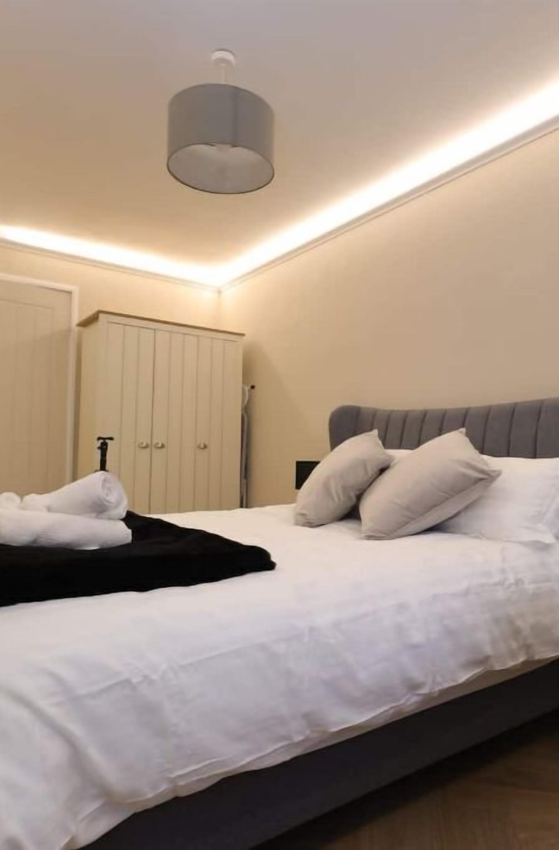 Cama bem arrumada com lençóis e almofadas brancos, cabeceira cinza e mesa de cabeceira com abajur.