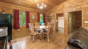 A wooden cabin interior with a dining table, chairs, a black couch, and a kitchen area.