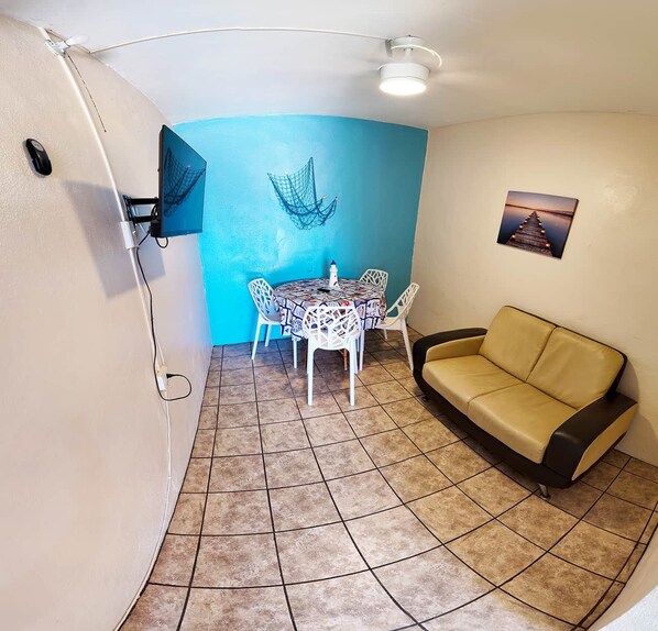 A small room with a tiled floor, a blue wall, a white table with chairs, a yellow couch, a mounted TV, and a picture on the wall.