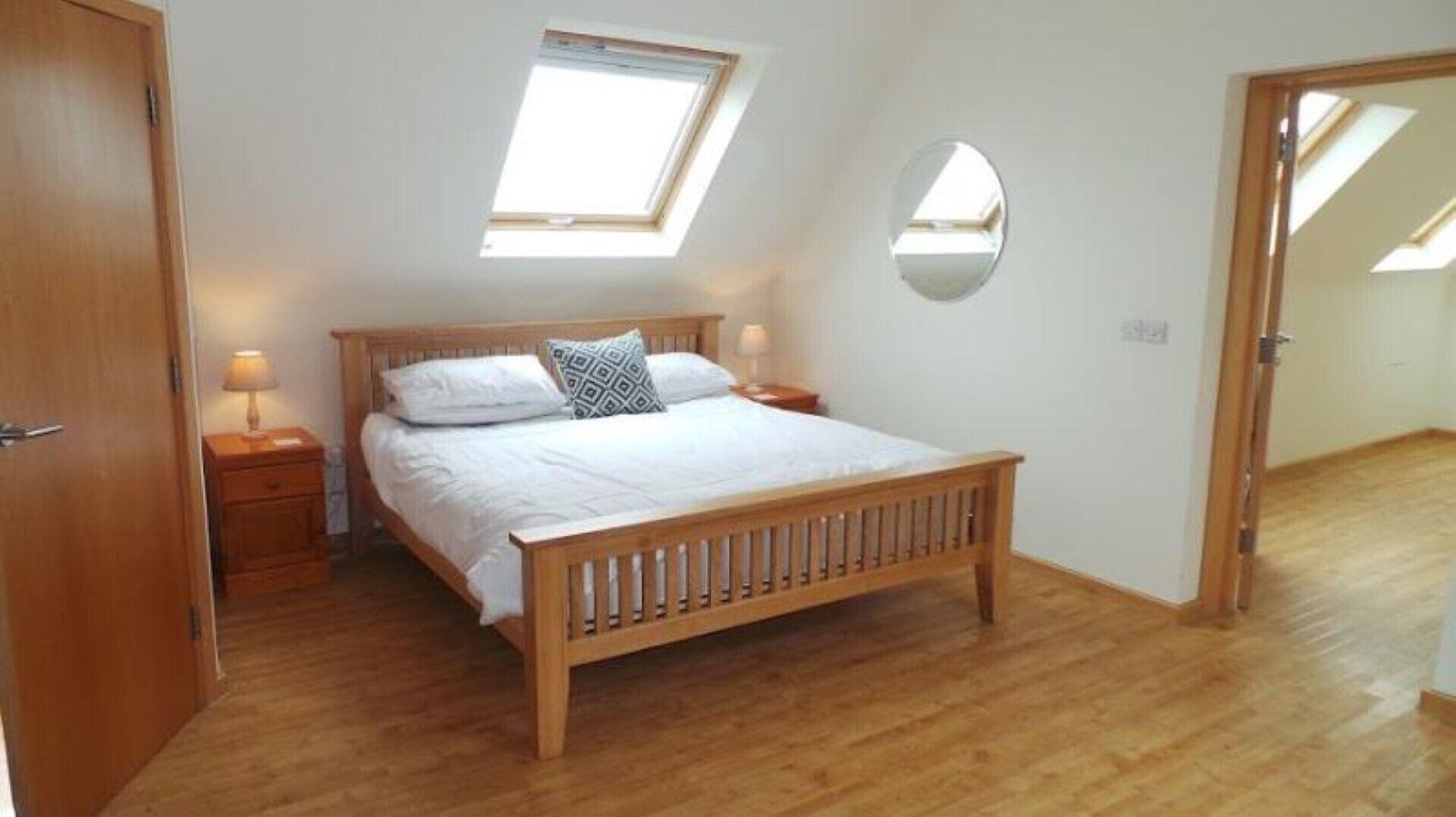 A bedroom with a wooden bed, a skylight, a round mirror, and wooden flooring.