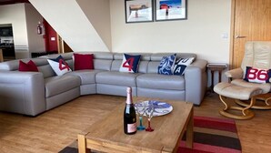 Ein modernes Wohnzimmer mit einem grauen Ecksofa, einem Holztisch und einem kleinen Beistelltisch. An der Wand hängen zwei gerahmte Bilder und im Hintergrund ist eine Holztür zu sehen.