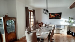 Un coin repas avec une longue table dressée pour six personnes, entourée de chaises. On y trouve, à gauche, un meuble avec des portes en verre, une fenêtre avec des rideaux et un tableau accroché au mur.