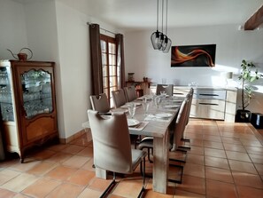 Een eethoek met een lange tafel, gedekt voor zes personen, omringd door stoelen. Links een kast met glazen deuren, een raam met gordijnen, en een schilderij aan de muur.