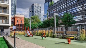 Un'area giochi con scivolo e triciclo, recintata da una staccionata e dotata di panchine, in un contesto urbano con palazzi alti.