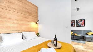 Camera d'albergo moderna con una testiera in legno, un letto con lenzuola bianche, una coperta gialla, una bottiglia di vino e un bicchiere su un vassoio rotondo in legno.