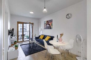 A modern living space with a sofa, dining table, and chairs, a small desk with a computer, and a large window.