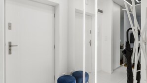 A modern hallway with white walls, a door, and a mirror reflecting a room with a black chair and a white ladder. - Comfortable studio on Twarda in Center (Warszawa)