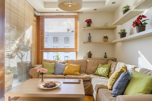 A modern living room with a beige sofa, a wooden coffee table with a plate of snacks, and a laptop. There are potted plants on shelves and a window with blinds.
