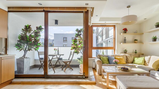 Una sala de estar moderna con un ventanal corredera que da a un balcón, un conjunto de sofás, una mesa de comedor y una pequeña área de cocina.
