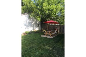 Relax in the serene backyard with a picnic table and umbrella, surrounded by lush greenery and a charming brick building in the distance.