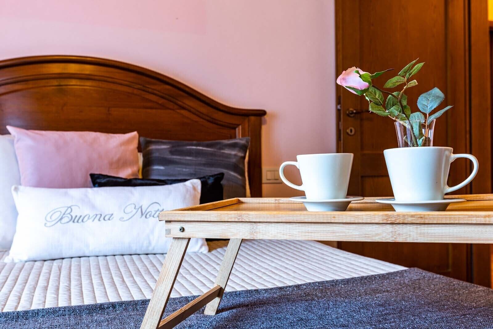 A neatly made bed with a headboard, a bedside table with two white cups and a vase of flowers, and a pillow with the text 'Buona Notte'.