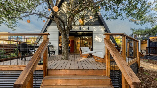 Une terrasse en bois avec un escalier menant à l’entrée d’un bâtiment, le tout entouré d’arbres et de mobilier d’extérieur.