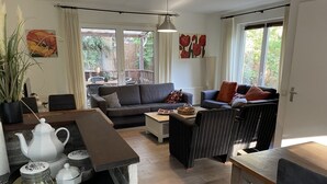 Un salon moderne comprenant un canapé, des fauteuils, une table basse et un coin repas avec une table et des chaises. Il y a une grande fenêtre avec des rideaux, une plante en pot et des œuvres d'art accrochées aux murs.