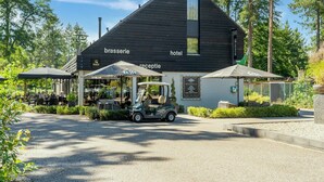 Il s’agit d’un bâtiment sur lequel est apposée une enseigne indiquant qu’il abrite un restaurant-brasserie et un hôtel. On y trouve des espaces de restauration en plein air avec des parasols, un véhicule de type voiturette de golf garé devant, et une allée gravillonnée.