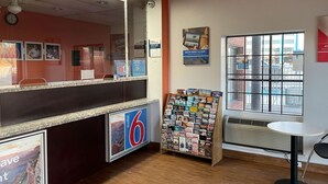 Un hall d’hôtel avec un comptoir d’accueil, un panneau d’affichage où sont accrochées diverses brochures, une horloge et une fenêtre donnant sur les bâtiments environnants.