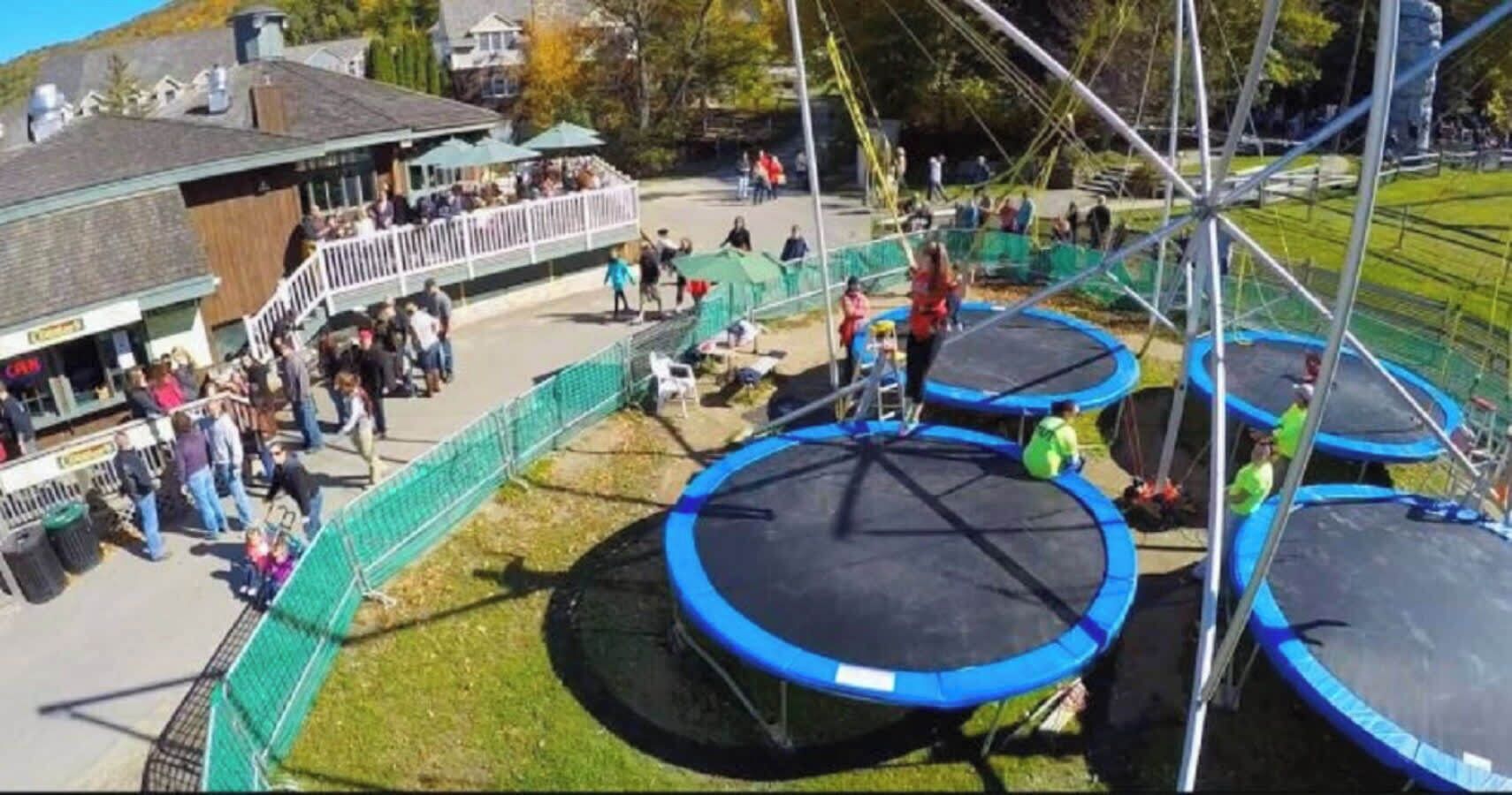 Trampoliinipuisto, jossa on useita trampoliineja ja turvaverkkoja, ja jonka ympärillä on aidattu alue, jossa katsojat voivat oleskella.