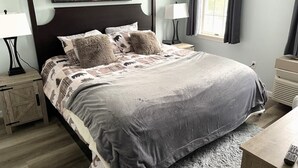 Un letto rifatto con una coperta grigia, due cuscini e una testiera. Si vede un comodino con una lampada, un tavolino in legno e un'opera d'arte appesa al muro.