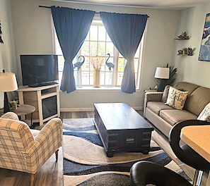 Una sala de estar con televisión, sofá, mesa de centro y un ventanal decorativo con cortinas.