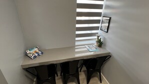 Un petit espace repas, à l’intérieur, avec une table grise et des chaises noires, une fenêtre avec des stores et une petite plante en pot posée sur la table.