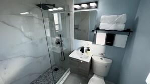 Une salle de bain moderne avec une douche à parois de verre, un lavabo avec un miroir au-dessus et des toilettes. Des serviettes et une serviette pour se maquiller sont suspendues à un porte-serviettes.