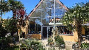 A modern building with large glass windows and a triangular roof, surrounded by tropical plants and a small garden area. - Villa indoor pool, hamman, sauna, jacuzzi, gym, billiards, baby (Plouezoc'h)