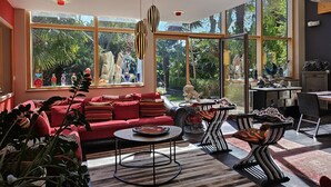 A spacious living room with a red sofa, a coffee table, and a dining area with a patterned rug. Large windows offer a view of greenery outside. - Villa indoor pool, hamman, sauna, jacuzzi, gym, billiards, baby (Plouezoc'h)