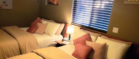 Un dormitorio con dos camas, cada una con cabecera, mesita de noche y lámpara, y una ventana con persianas.