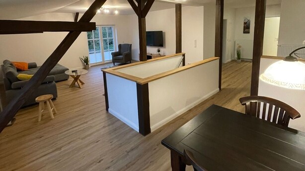 A spacious living area with a wooden dining table, a sofa, and a small stool. There is a kitchen area with a counter and a television. - New vacation apartment with roof terrace (Bünsdorf)