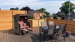 A wooden deck with a grill, a table with chairs, and a bench with a plaid cushion. - New vacation apartment with roof terrace (Bünsdorf)