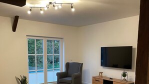 A spacious living room with hardwood flooring, a grey armchair, a wooden TV stand, and a large window offering a view of greenery. - New vacation apartment with roof terrace (Bünsdorf)